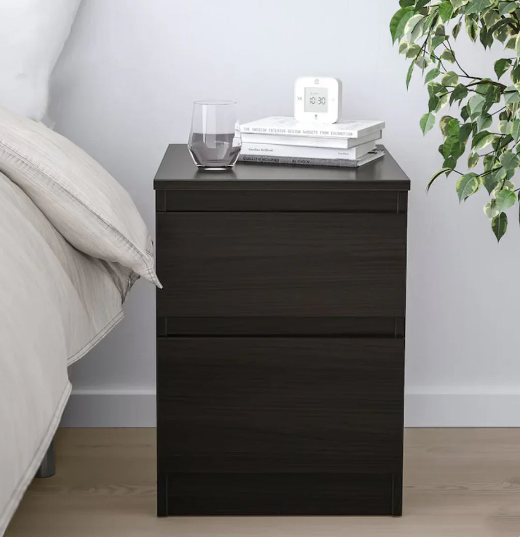 Black nightstand with books, a glass, and a clock next to a bed with a plant in the corner.